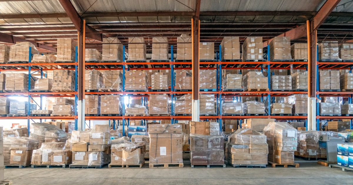 A wall of pallet racks as a row of pallets lined up in front of it on the ground. The pallets have boxes that are plastic wrapped.