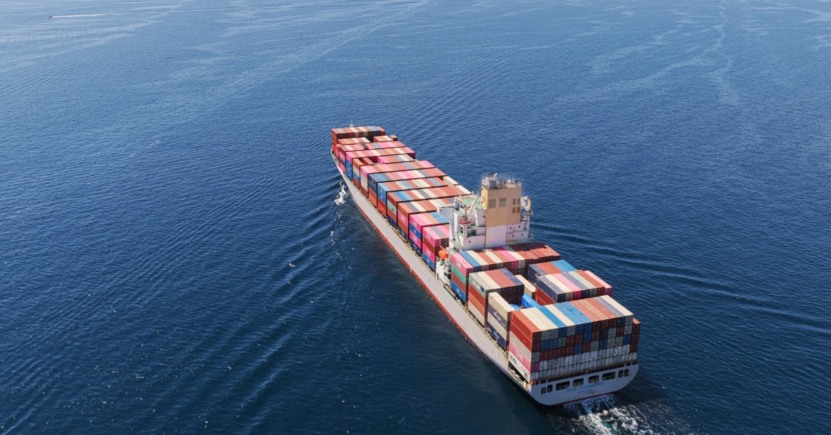 A container ship is sailing away from the camera. It is heading out into open ocean. The sun is also out.