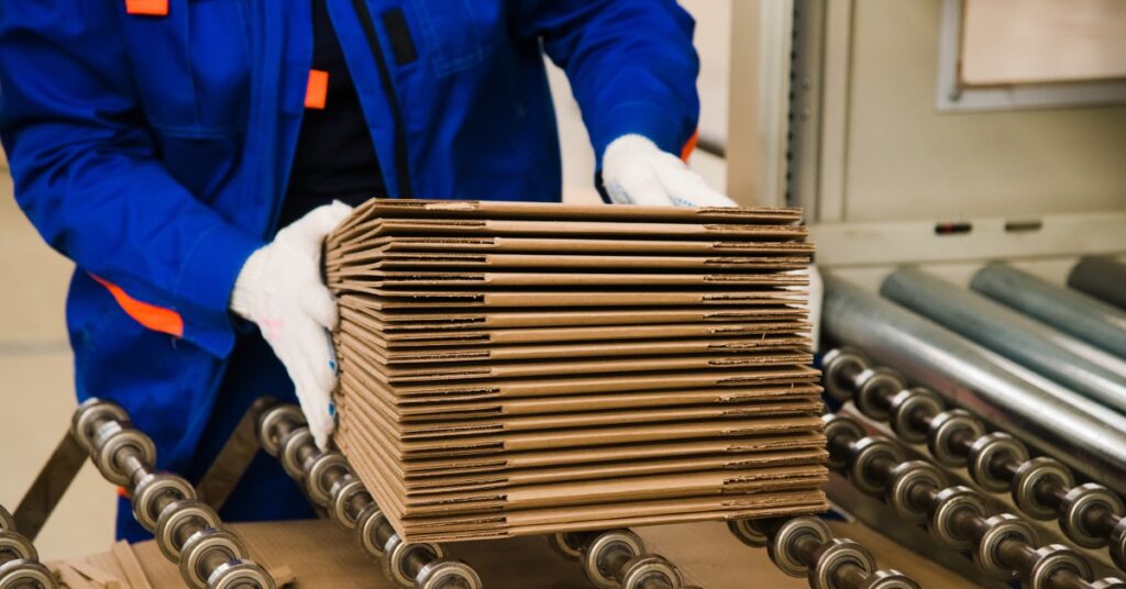A person in a blue jacket and wearing gloves is holding a stack of unfolded cardboard in their hands.