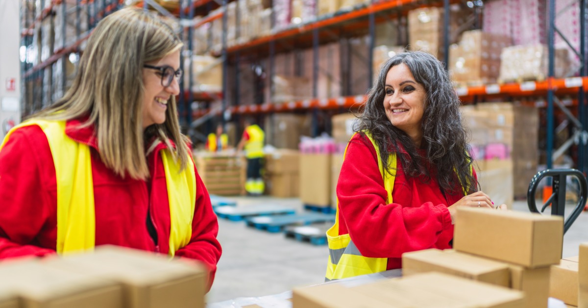 Two women, both of whom are wearing red shirts and yellow vests, are packing boxes in a warehouse.