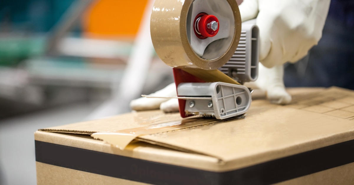 A person is using a tape gun to seal a cardboard box. The person is wearing white gloves.