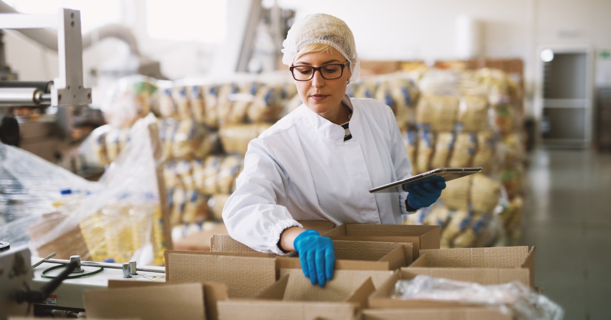 A woman in a white coat and a hairnet is standing over a table full of cardboard boxes, observing them.
