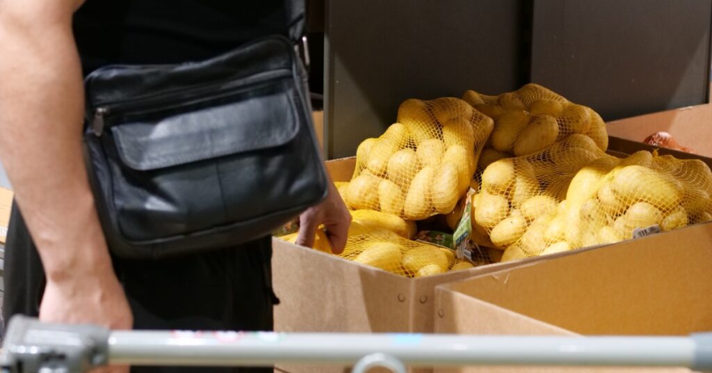 A person wearing a black satchel is standing next to a large cardboard bin full of potatoes.